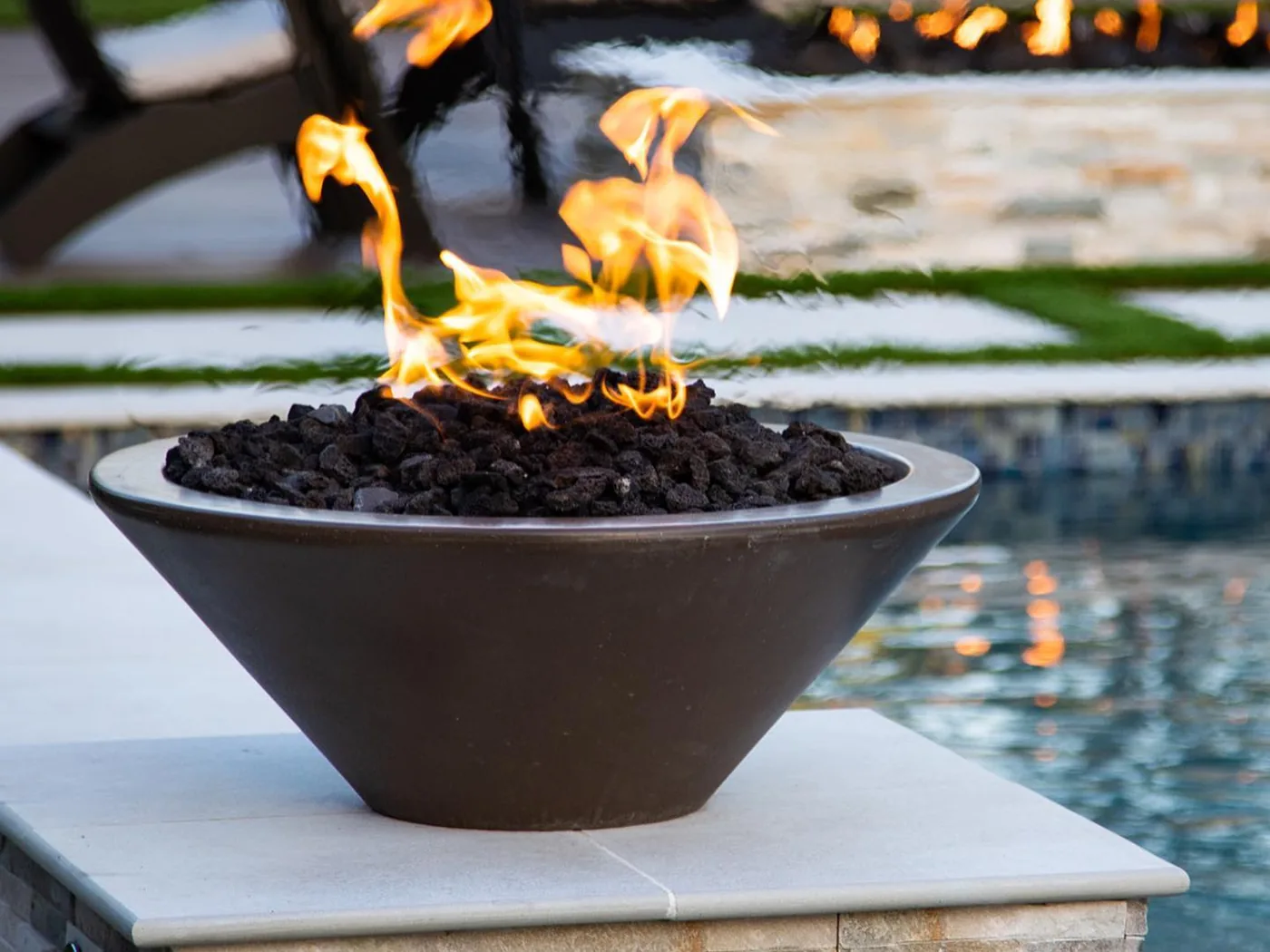 A The Outdoor Plus Cazo fire bowl burning on a stacked-stone pilaster at the edge of a pool